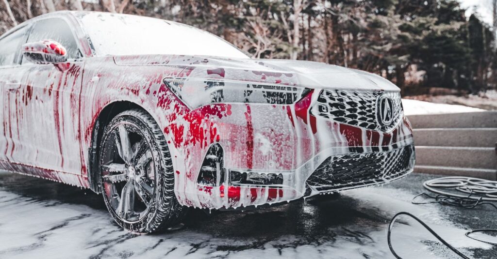 A vibrant red car covered in soapy foam during a thorough outdoor wash under daylight.