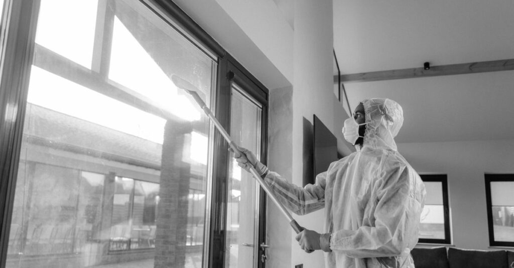 Person in protective coveralls sanitizing a window indoors with daylight.