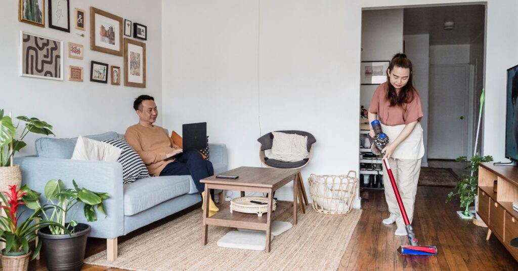 A man working on a laptop while a woman vacuums the living room, showcasing modern lifestyle balance.